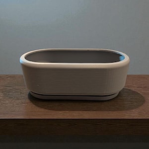 Puede incluir: Una jardinera rectangular blanquecina con interior gris se encuentra sobre una superficie de madera. La jardinera tiene un exterior texturizado y un borde redondeado. El fondo es de color gris claro s&oacute;lido.