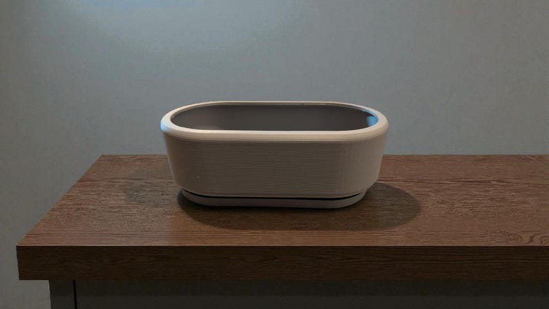Puede incluir: Una jardinera rectangular blanquecina con interior gris se encuentra sobre una superficie de madera. La jardinera tiene un exterior texturizado y un borde redondeado. El fondo es de color gris claro s&oacute;lido.