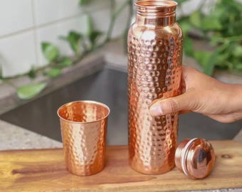 May include: A copper water bottle with a hammered texture, a matching cup, and a lid. The bottle is held by a person. The items are on a wooden surface.