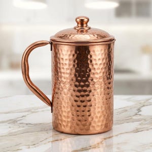 Puede incluir: Una jarra de color cobre con textura martillada, asa y tapa. La jarra es cilíndrica y se encuentra sobre una superficie de mármol. El asa es curvada y la tapa tiene un pequeño pomo. El diseño general es simple y elegante.