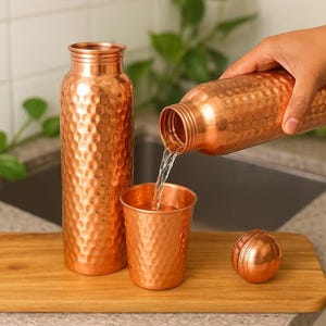 K&ouml;nnte beinhalten: Eine Kupferwasserflasche, ein Becher und ein Deckel werden auf einem Holztablett pr&auml;sentiert. Wasser wird aus der Flasche in den Becher gegossen. Die Kupferartikel haben eine strukturierte, gew&ouml;lbte Oberfl&auml;che. Die Flasche ist etwa 25 cm hoch.