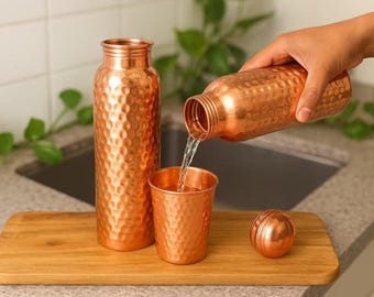 May include: A copper water bottle, cup, and bottle are displayed on a wooden tray. Water is being poured from the bottle into the cup. The copper items have a textured, dimpled surface. The bottle is approximately 25 cm tall.