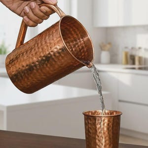 May include: A hammered copper pitcher with a curved handle pouring clear water into a matching hammered copper tumbler. The metallic surface of both vessels features a textured, dimpled finish that reflects light across the warm, reddish-brown exterior.