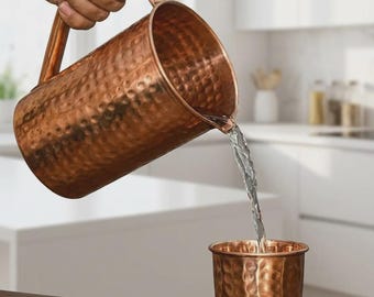 May include: A hammered copper pitcher with a curved handle pouring clear water into a matching hammered copper tumbler. The metallic surface of both vessels features a textured, dimpled finish that reflects light across the warm, reddish-brown exterior.