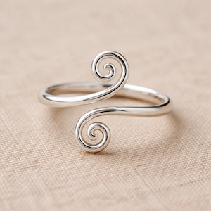 Könnte beinhalten: Ein silberner Ring mit einem einzigartigen Design. Der Ring hat zwei wirbelnde Spiralakzente, die ein offenes, verstellbares Design bilden. Der Ring wird auf einer neutral gefärbten Oberfläche gezeigt.