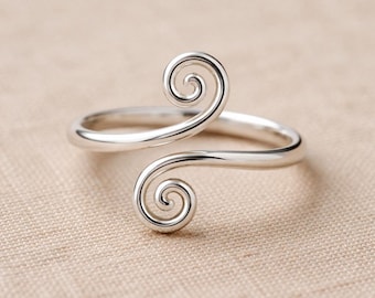 May include: A silver ring with a distinctive design. The ring has two swirling spiral features, forming an open, adjustable design. The ring is displayed on a neutral-toned surface.