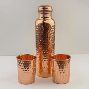 Puede incluir: Una botella de agua de cobre con textura martillada, acompa&ntilde;ada de dos vasos de cobre a juego. La botella tiene una tapa de rosca. El juego se exhibe sobre una superficie blanca.