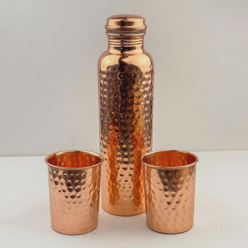 Puede incluir: Una botella de agua de cobre con textura martillada, acompa&ntilde;ada de dos vasos de cobre a juego. La botella tiene una tapa de rosca. El juego se exhibe sobre una superficie blanca.