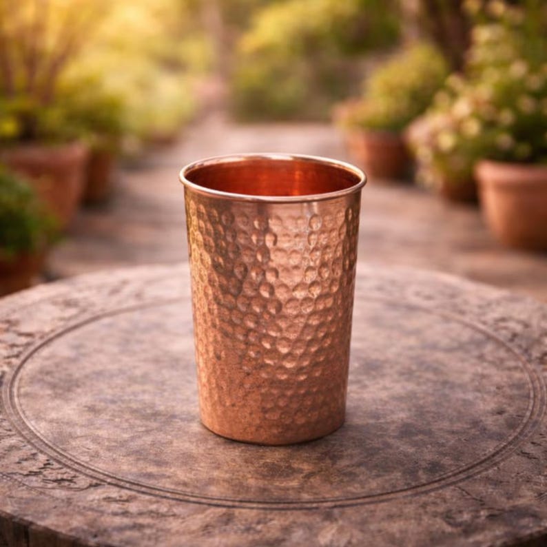 K&ouml;nnte beinhalten: Ein kupferfarbenes Trinkglas mit gehammerter Textur. Das Glas ist zylindrisch mit einem leicht ausgestellten Rand. Das Glas steht auf einem runden, steinartigen Tisch. Der Hintergrund ist verschwommen und zeigt eine Gartenumgebung.