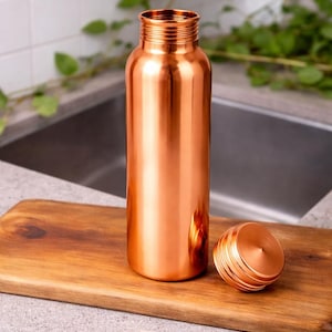Puede incluir: Una botella de agua de color cobre con una tapa de rosca a juego, colocada sobre una tabla de cortar de madera. La botella tiene forma cil&iacute;ndrica y un acabado brillante. La tapa est&aacute; al lado de la botella. El fondo incluye un fregadero y vegetaci&oacute;n.