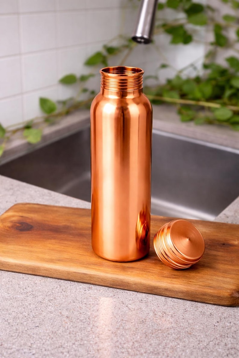 Puede incluir: Una botella de agua de color cobre con una tapa de rosca a juego, colocada sobre una tabla de cortar de madera. La botella tiene forma cil&iacute;ndrica y un acabado brillante. La tapa est&aacute; al lado de la botella. El fondo incluye un fregadero y vegetaci&oacute;n.