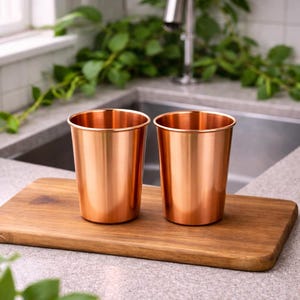Puede incluir: Dos vasos de metal color cobre sobre una tabla de cortar de madera. Los vasos tienen una superficie brillante y reflectante y una forma cil&iacute;ndrica simple. La tabla de cortar es rectangular con bordes redondeados. El fondo incluye un fregadero de cocina y vegetaci&oacute;n.
