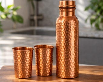 May include: A copper water bottle and two matching cups are displayed on a wooden surface. The bottle has a textured, hammered finish and a screw-on cap. The cups have a similar hammered texture. The items are in a kitchen setting.