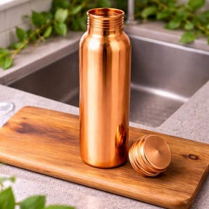 May include: A polished copper water bottle with a smooth, cylindrical body and a threaded neck. The matching circular copper cap is placed beside the bottle on a wooden surface. The metal surface reflects light, showing a warm, metallic orange hue.
