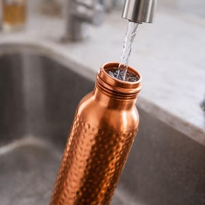 K&ouml;nnte beinhalten: Eine Kupfer-Wasserflasche wird mit Wasser aus einem Wasserhahn aus Edelstahl gef&uuml;llt. Die Flasche hat eine geh&auml;mmerten Textur und eine weite &Ouml;ffnung. Der Hintergrund umfasst ein Edelstahlbecken und eine verschwommene K&uuml;che.