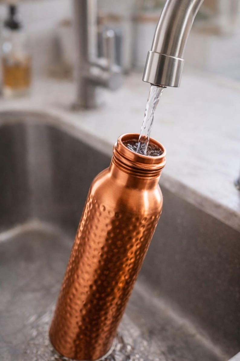 K&ouml;nnte beinhalten: Eine Kupfer-Wasserflasche wird mit Wasser aus einem Wasserhahn aus Edelstahl gef&uuml;llt. Die Flasche hat eine geh&auml;mmerten Textur und eine weite &Ouml;ffnung. Der Hintergrund umfasst ein Edelstahlbecken und eine verschwommene K&uuml;che.