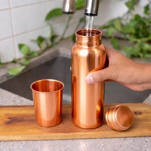 Puede incluir: Una botella de agua de cobre se llena con agua del grifo. Una taza de cobre a juego y la tapa de la botella est&aacute;n sobre una tabla de madera. La botella y la taza tienen una superficie brillante y reflectante.