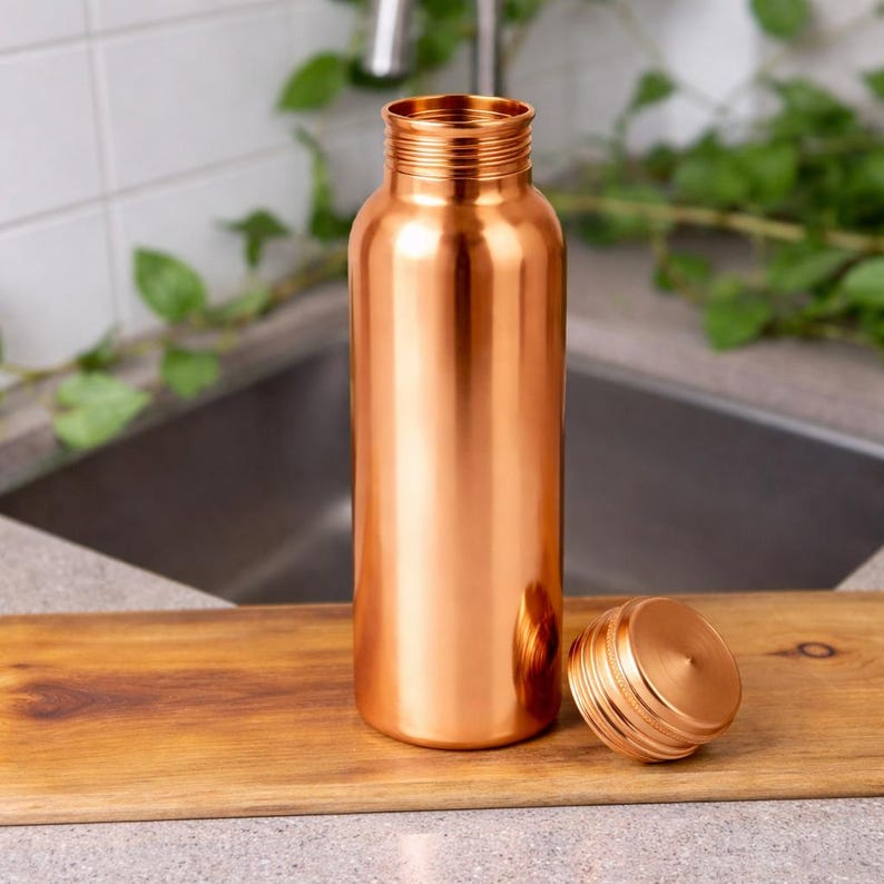 Puede incluir: Una botella de agua de color cobre con una tapa de rosca a juego. La botella es cil&iacute;ndrica con una boca ancha y se encuentra sobre una tabla de cortar de madera. La tapa est&aacute; al lado de la botella. El fondo incluye un fregadero y vegetaci&oacute;n.