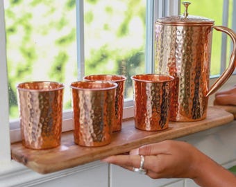 May include: A set of three hammered copper tumblers and a matching hammered copper pitcher with a curved handle and lid. The metallic drinkware set features a textured, dimpled surface that reflects light, arranged on a wooden ledge.