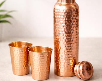May include: A copper water bottle with a hammered texture, alongside two small copper cups and a lid. The bottle is tall and cylindrical, and the cups are small and round. The set is displayed on a light-coloured surface.