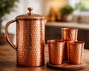 May include: A copper jug with a hammered finish, a lid, and a handle, alongside three copper cups and a wooden coaster. The jug and cups have a shiny surface. The set is on a wooden table.