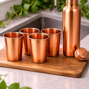 Puede incluir: Una botella de agua de color cobre con una tapa a juego y cuatro tazas de cobre est&aacute;n sobre una tabla de cortar de madera. La botella y las tazas son cil&iacute;ndricas. Los art&iacute;culos est&aacute;n en un entorno de cocina.
