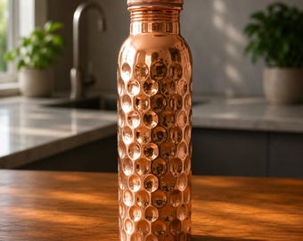 May include: A cylindrical copper water bottle featuring a distinct hammered texture across its entire surface. The bottle has a narrow neck and a matching metallic screw-top lid, reflecting light with a warm, polished, and reflective finish.