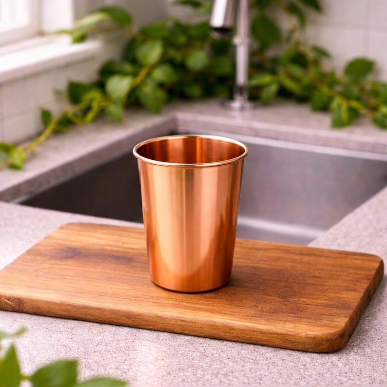 Puede incluir: Un vaso de color cobre se encuentra sobre una tabla de cortar de madera. La taza tiene una superficie lisa y reflectante y un borde redondeado. La tabla de cortar es rectangular con bordes redondeados. El fondo incluye un fregadero de cocina y vegetaci&oacute;n.