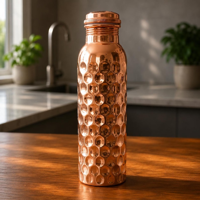 May include: A cylindrical copper water bottle featuring a distinct hammered texture across its entire surface. The bottle has a narrow neck and a matching metallic screw-top lid, reflecting light with a warm, polished, and reflective finish.