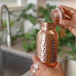 Könnte beinhalten: Eine Kupfer-Wasserflasche mit einer strukturierten, gewölbten Oberfläche. Die Flasche ist zylindrisch mit einem Schraubverschluss. Der Deckel wird gerade entfernt, wodurch die Öffnung freigelegt wird. Die Flasche wird von Händen gehalten, die Ringe tragen. Der Hintergrund umfasst ein Waschbecken und Grün.