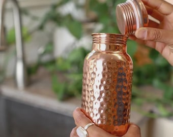 May include: A copper water bottle with a textured, dimpled surface. The bottle is cylindrical with a screw-on lid. The lid is being removed, revealing the opening. The bottle is held by hands wearing rings. The background includes a sink and greenery.
