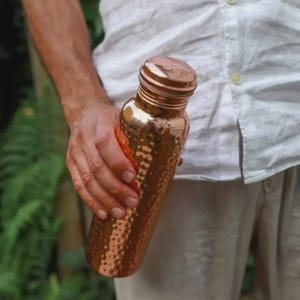 Puede incluir: Una botella de agua de cobre con textura martillada. La botella la sostiene una persona que lleva una camisa blanca. La botella tiene una tapa de rosca y un acabado met&aacute;lico brillante. La botella mide aproximadamente 25 cm de alto.