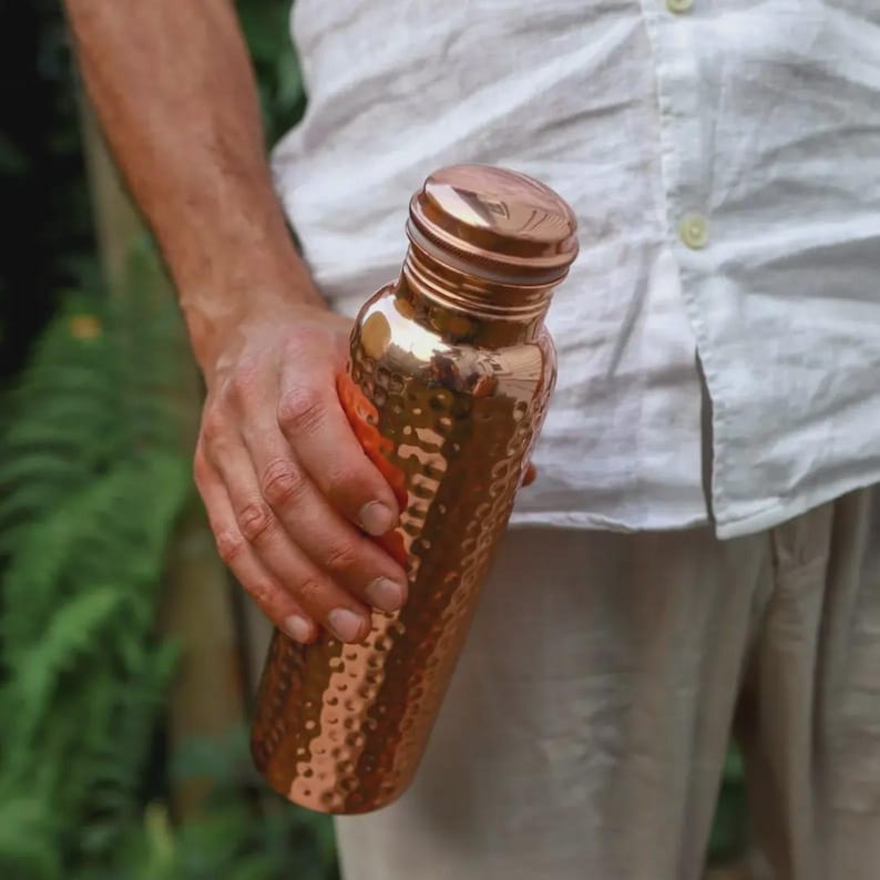 Puede incluir: Una botella de agua de cobre con textura martillada. La botella la sostiene una persona que lleva una camisa blanca. La botella tiene una tapa de rosca y un acabado met&aacute;lico brillante. La botella mide aproximadamente 25 cm de alto.