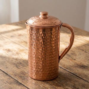 May include: A copper jug with a hammered texture, a lid, and a handle. The jug is cylindrical and sits on a wooden surface. The copper has a warm, reddish-brown hue.