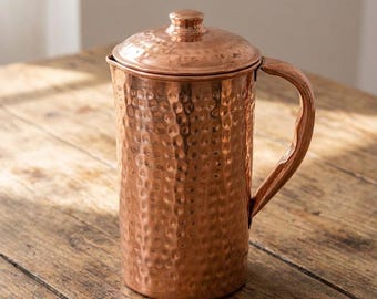 May include: A copper pitcher with a hammered texture and a matching lid. The pitcher has a curved handle and is sitting on a wooden surface. The copper color is a warm, reddish-brown hue.