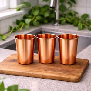 Puede incluir: Tres vasos de metal color cobre est&aacute;n colocados sobre una tabla de cortar de madera. Los vasos tienen un acabado brillante y una forma cil&iacute;ndrica simple. La tabla es rectangular y tiene vetas de madera natural.