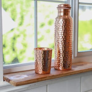 Puede incluir: Una botella de agua de cobre y un vaso a juego est&aacute;n sobre una repisa de madera. Ambos art&iacute;culos tienen una textura martillada y un acabado met&aacute;lico brillante. La botella es alta y cil&iacute;ndrica con una tapa de rosca. El vaso es una taza corta y recta.