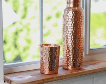 May include: A copper water bottle and a matching tumbler are displayed on a wooden shelf. Both items have a hammered texture and a shiny, metallic finish. The bottle is tall and cylindrical with a screw-on lid. The tumbler is a short, straight-sided cup.