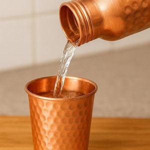 K&ouml;nnte beinhalten: Eine Kupferwasserflasche gie&szlig;t Wasser in einen passenden Kupferbecher. Beide Artikel haben eine geh&auml;mmerten Textur. Der Wasserstrahl ist klar und der Hintergrund ist neutral.