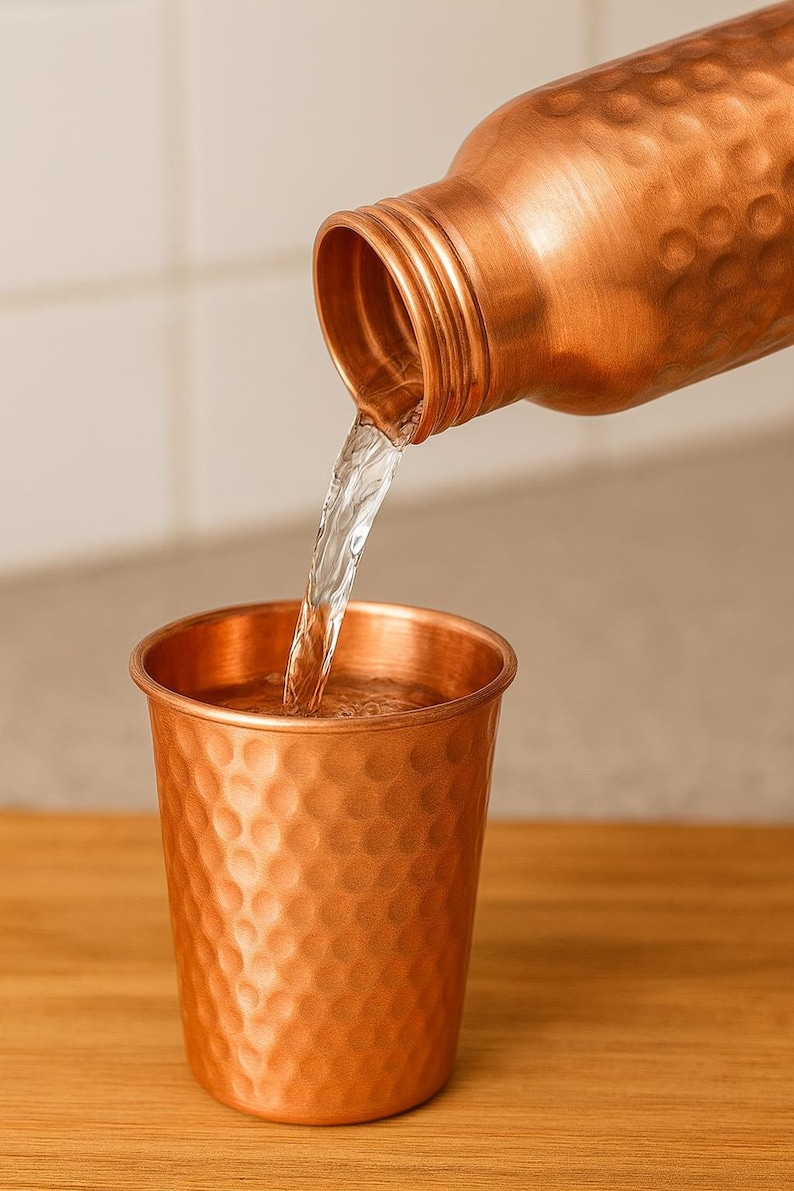 K&ouml;nnte beinhalten: Eine Kupferwasserflasche gie&szlig;t Wasser in einen passenden Kupferbecher. Beide Artikel haben eine geh&auml;mmerten Textur. Der Wasserstrahl ist klar und der Hintergrund ist neutral.