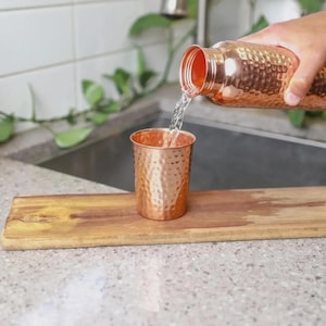 Puede incluir: Una botella de agua de cobre vierte agua en una taza de cobre a juego. Ambos art&iacute;culos tienen una textura martillada. La taza se encuentra sobre una tabla de madera. El fondo incluye un fregadero y vegetaci&oacute;n.