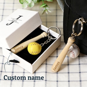 May include: A baseball-themed keychain and a miniature baseball bat and ball set in a white gift box. The keychain features a small wooden bat with "James" engraved on it, and a baseball charm. The gift box also has "James" on it.