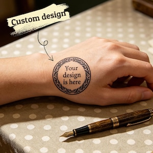 May include: A temporary tattoo with a circular design on a wrist. The tattoo features a decorative border and the text "Your design is here." The words "Custom design" are above the tattoo. A brown and gold fountain pen rests nearby.