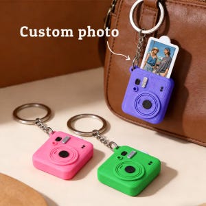 Puede incluir: Tres llaveros con forma de cámara en color morado, rosa y verde, cada uno con una anilla y cadena de metal plateado. El llavero morado muestra una pequeña fotografía rectangular con dos personas. El texto Custom photo es visible.