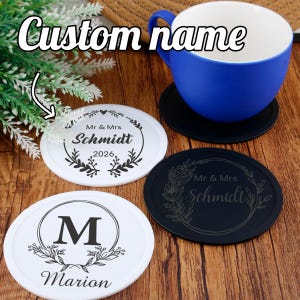 Pode incluir: Uma caneca azul está sobre um porta-copos preto, com três porta-copos personalizados abaixo. Dois porta-copos brancos apresentam desenhos florais e os nomes "Schmidt" e "Marion". Um porta-copos preto também exibe "Mr & Mrs Schmidt". O texto "Custom name" está acima.