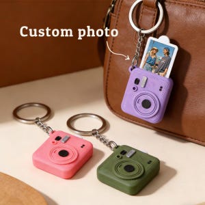 May include: Keychains in pink, purple, and green, each shaped like a retro camera. The purple keychain holds a small photo. The text "Custom photo" is visible in the image. The keychains are attached to silver keyrings.