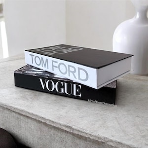May include: Two hardback books stacked on a light grey surface. The top book is black with "TOM FORD" in silver lettering. The bottom book is black with "VOGUE" in white lettering. A white vase is in the background.