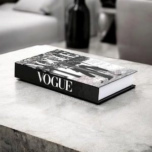 May include: A black and white decorative book with the word "VOGUE" in white lettering on the spine. The book's cover features a black and white photograph. The book is resting on a gray surface.