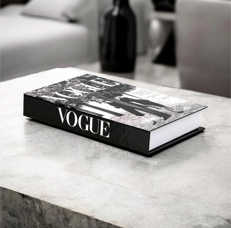 May include: A black and white decorative book with the word "VOGUE" in white lettering on the spine. The book's cover features a black and white photograph. The book is resting on a gray surface.