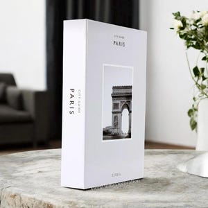 K&ouml;nnte beinhalten: Eine wei&szlig;e rechteckige Box mit den Worten "CITY GUIDE PARIS" auf der Vorder- und Seite aufgedruckt. Auf der Vorderseite befindet sich ein Schwarz-Wei&szlig;-Foto des Arc de Triomphe. Das Wort "CEREAL" ist unten aufgedruckt.